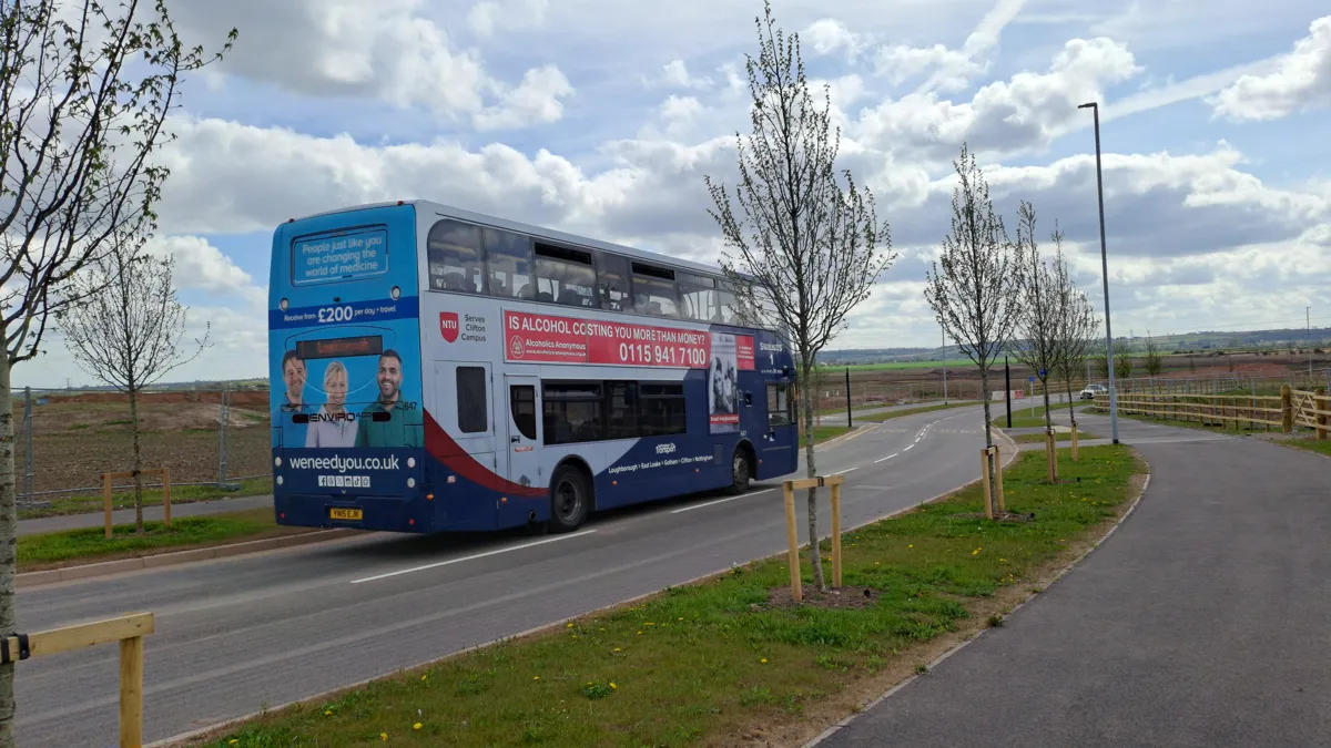 A Nottingham City Transport double-decker bus on Fairham Way, with young street trees and open land under development on either side