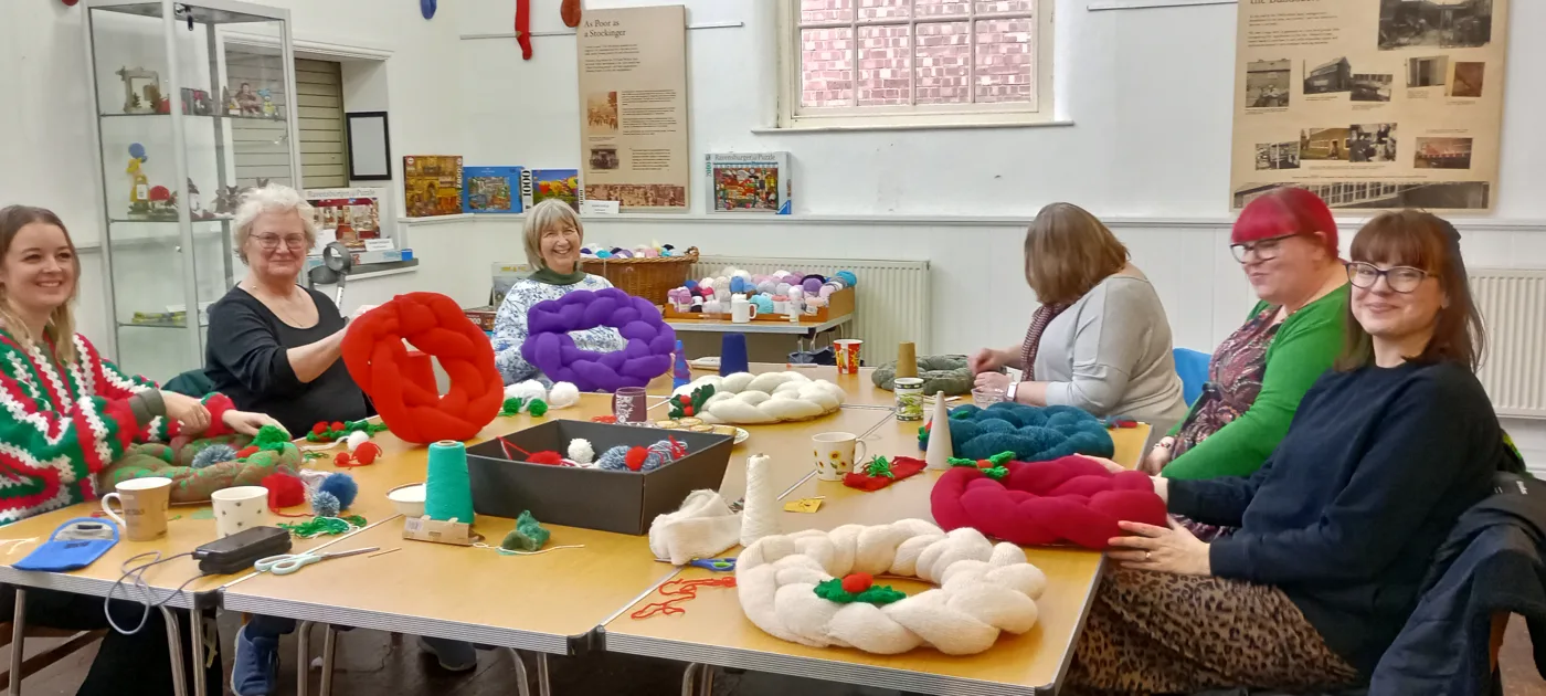 A group of people seated around tables with yarn and partly made knitted wreaths in white, red and purple