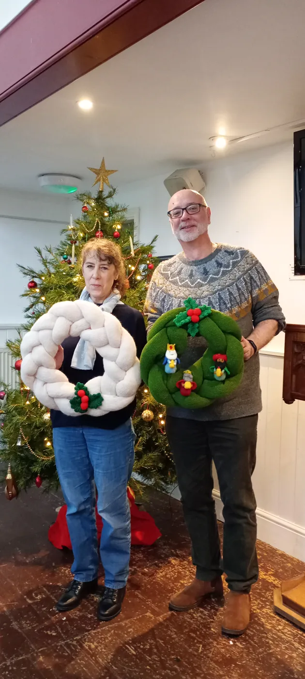 Two people standing by a Christmas tree, each holding a knitted wreath, one white and one green with small knitted figures