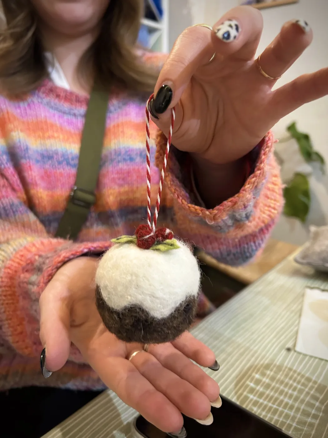 A hand holding up a needle-felted Christmas pudding ornament on a red and white string