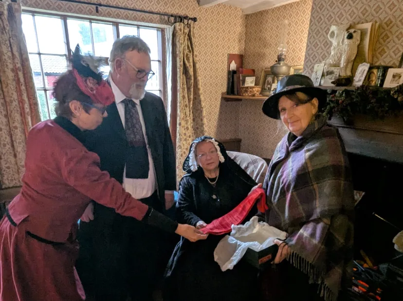 People in Victorian dress in a cottage parlour, holding up knitted stockings in red and white