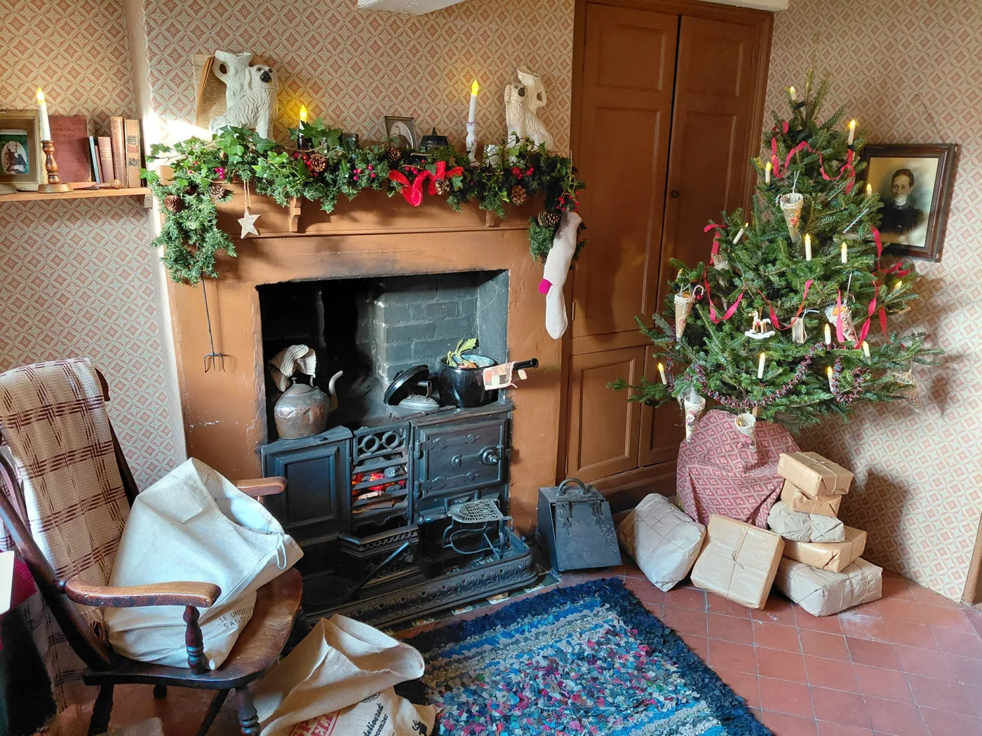 A cottage parlour with a black range, a mantelpiece hung with greenery, a candle-lit Christmas tree and wrapped parcels