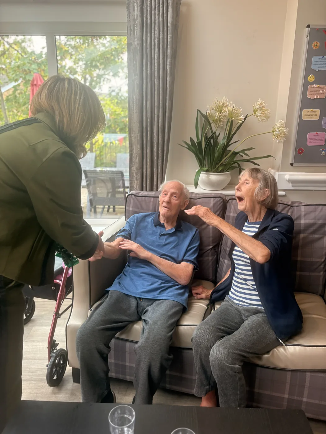 Lilian Greenwood MP shaking hands with a man in a blue polo shirt sitting on a sofa beside a woman in a striped top, in a lounge at Wilford View